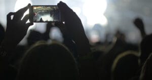 Hands in the crowd holding smart phone and shooting musical performance. Bright illumination on the stage